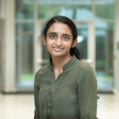 Portrait of Arya Patel smiling gently while standing indoors in a softly lit hallway with large windows in the background.