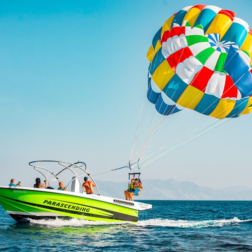 A green parasailing boat with people on board pulls a colorful parachute with two people ascending over the ocean.
