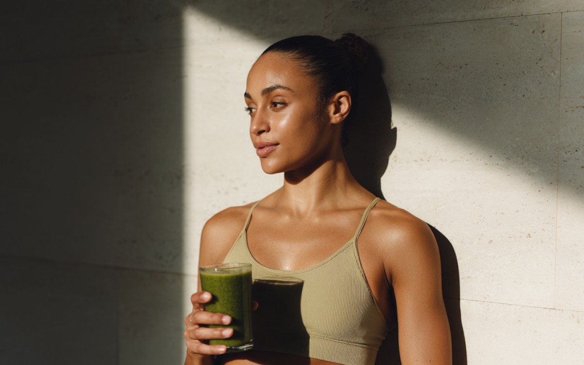 Woman in a tan sports top holds a green smoothie. She stands against a sunlit stone wall, conveying a calm, health-focused atmosphere.