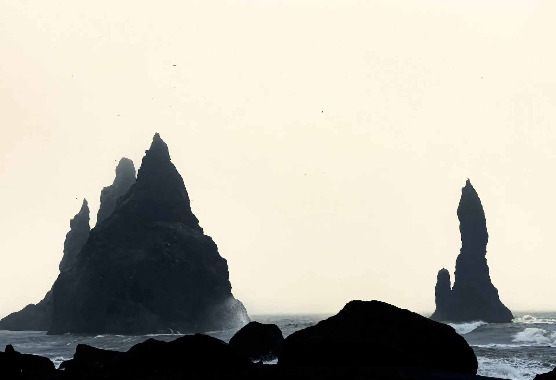 A white sky behind dark rock formations in the sea.