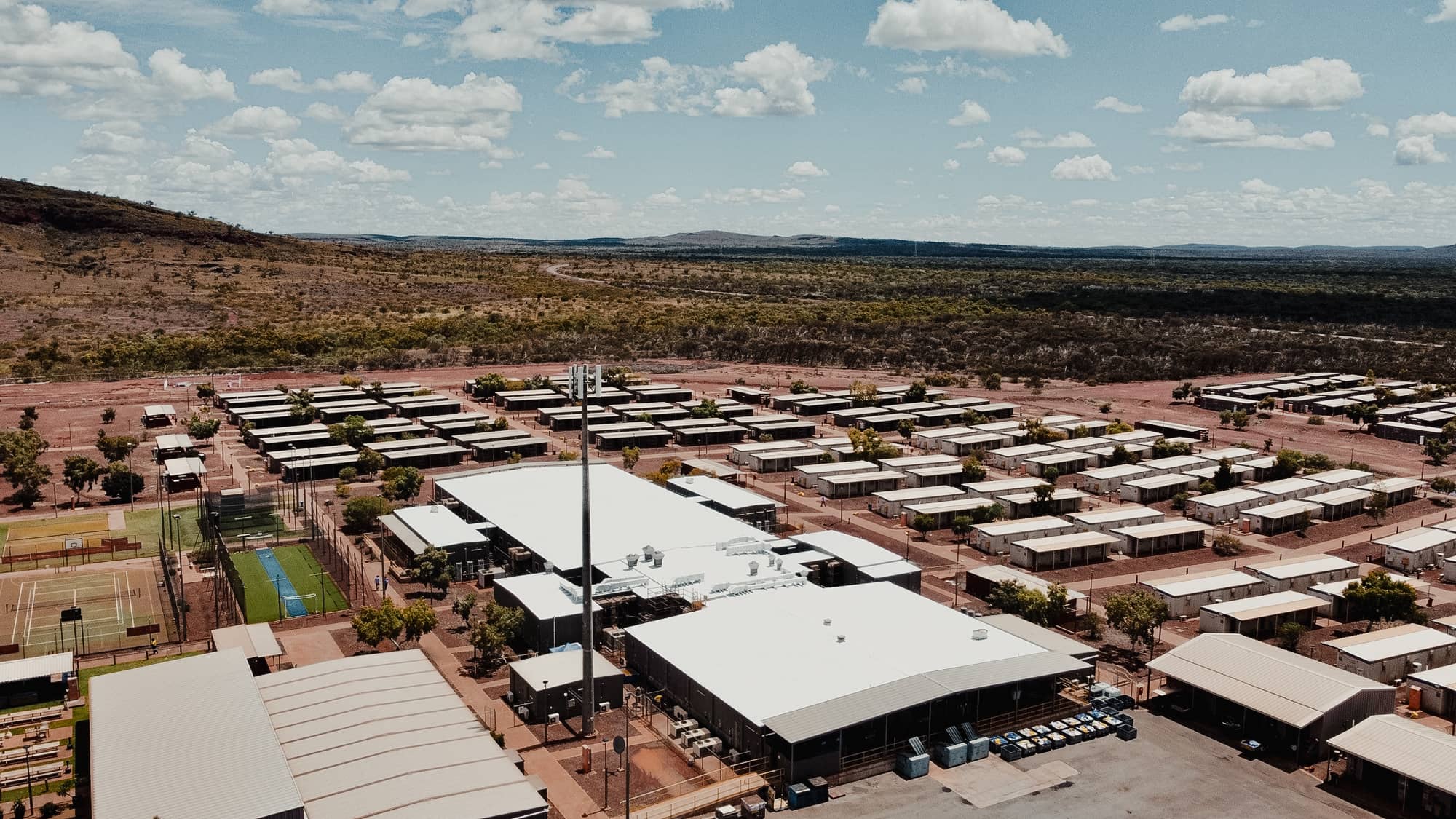 Aerial view of a remote mining camp with rows of prefabricated accommodation buildings, central facilities, and sports courts in a rugged Australian landscape.