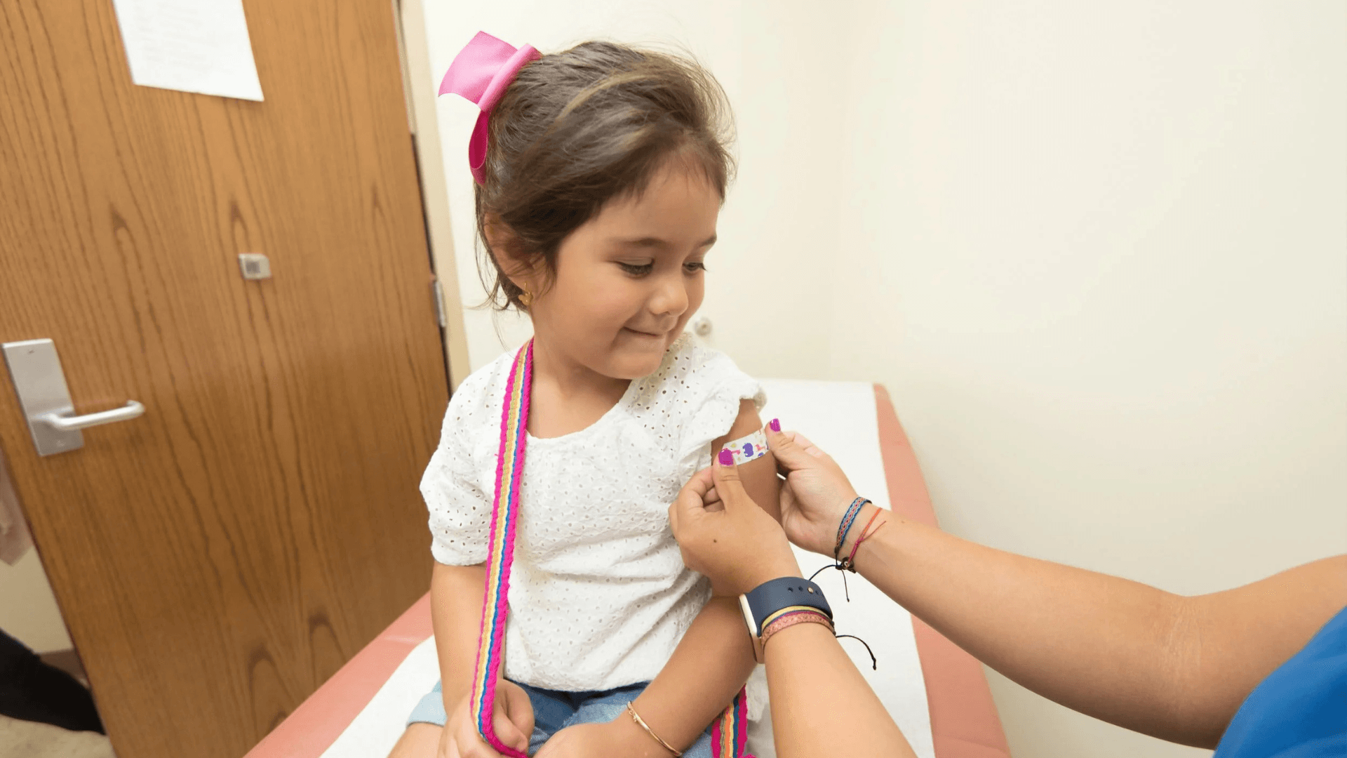 Alt tag: Young child sitting on an exam table while a caregiver applies a bandage to their upper arm during a medical visit