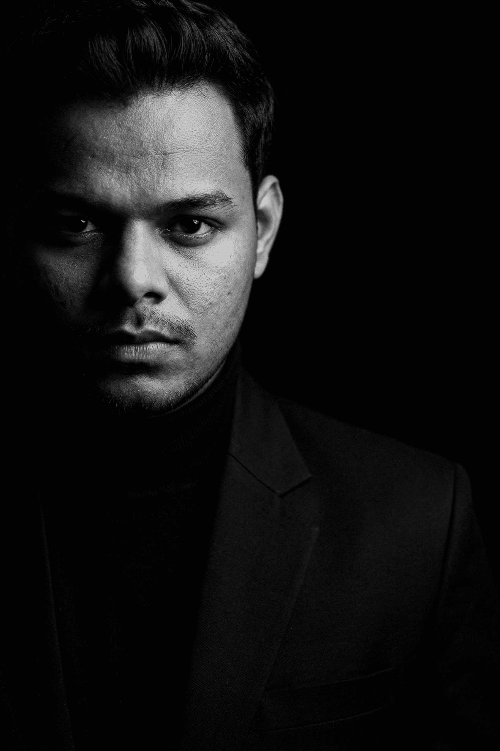 Smiling man with neat dark hair, glasses, black turtleneck, and gray blazer.