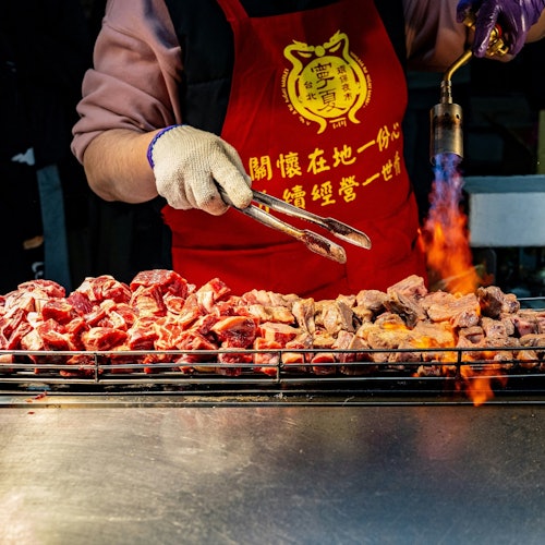 一位穿着印有黄色文字红色围裙的人，使用钳子和喷火枪，烤制串烧肉。