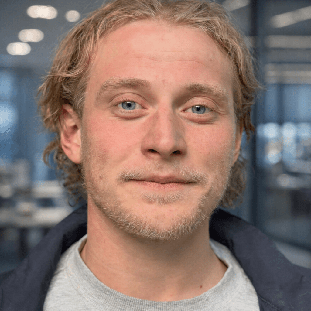 A young person with curly hair smiles at the camera while sitting in front of a laptop against a dark background.