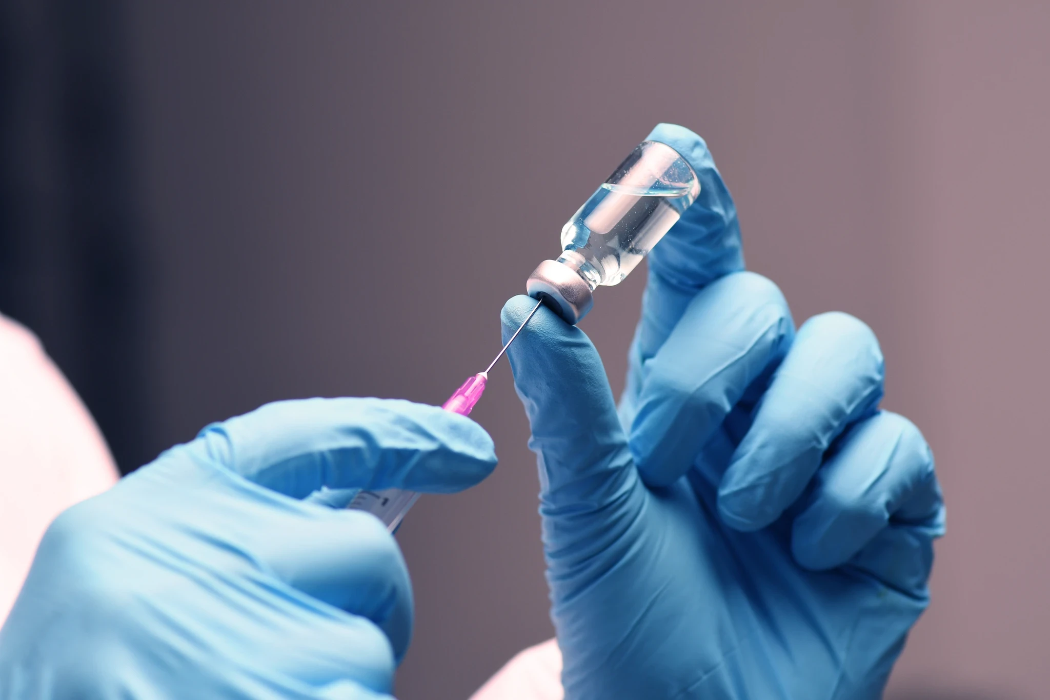 Gloved hands drawing liquid from a vial into a syringe with a pink needle hub.