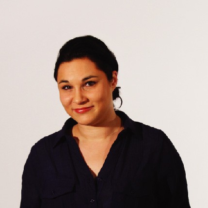 A woman with short, dark hair pulled back from her face wears a dark navy or black button-up shirt against a plain off-white background. She has dark eyes, a straight nose, and is wearing red lipstick. Her expression is neutral, and she looks directly at the camera.