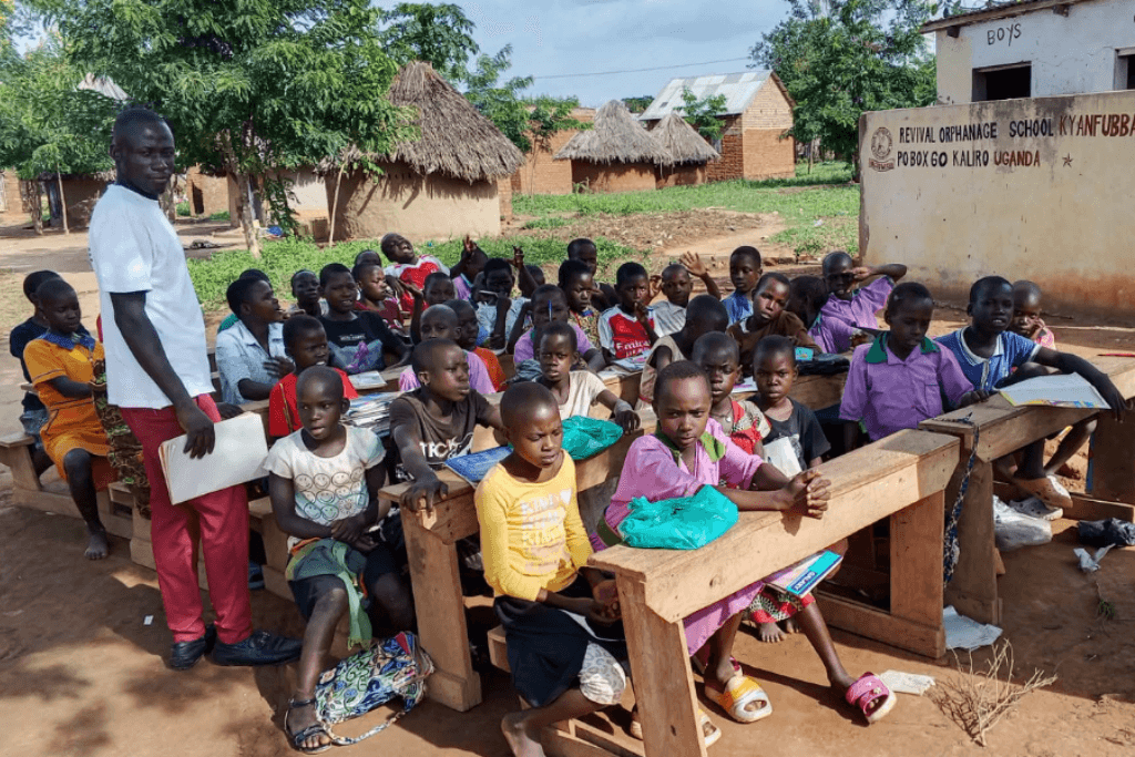 A teacher stands beside students seated outdoors at wooden desks, with school buildings and trees in the background. The setting feels collaborative and open.