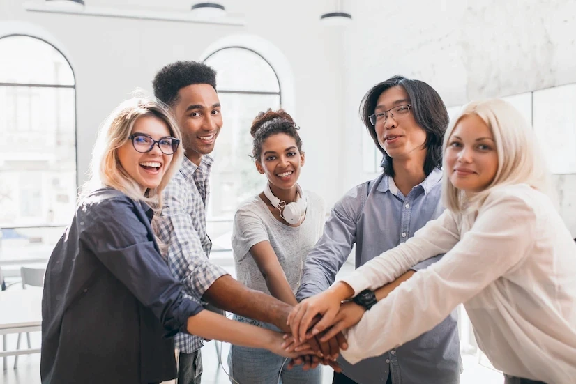 A imagem mostra um grupo de cinco pessoas sorridentes em um ambiente claro e iluminado, reunidas com as mãos unidas no centro, em um gesto de união e trabalho em equipe. Todos estão visivelmente alegres e entusiasmados. O grupo é diverso, com pessoas de diferentes etnias e estilos. Uma das pessoas usa óculos e outra está com fones de ouvido ao redor do pescoço, sugerindo um ambiente moderno e descontraído, possivelmente de trabalho colaborativo ou educacional. Ao fundo, grandes janelas permitem a entrada de luz natural, destacando o clima positivo e a sensação de união entre o grupo.