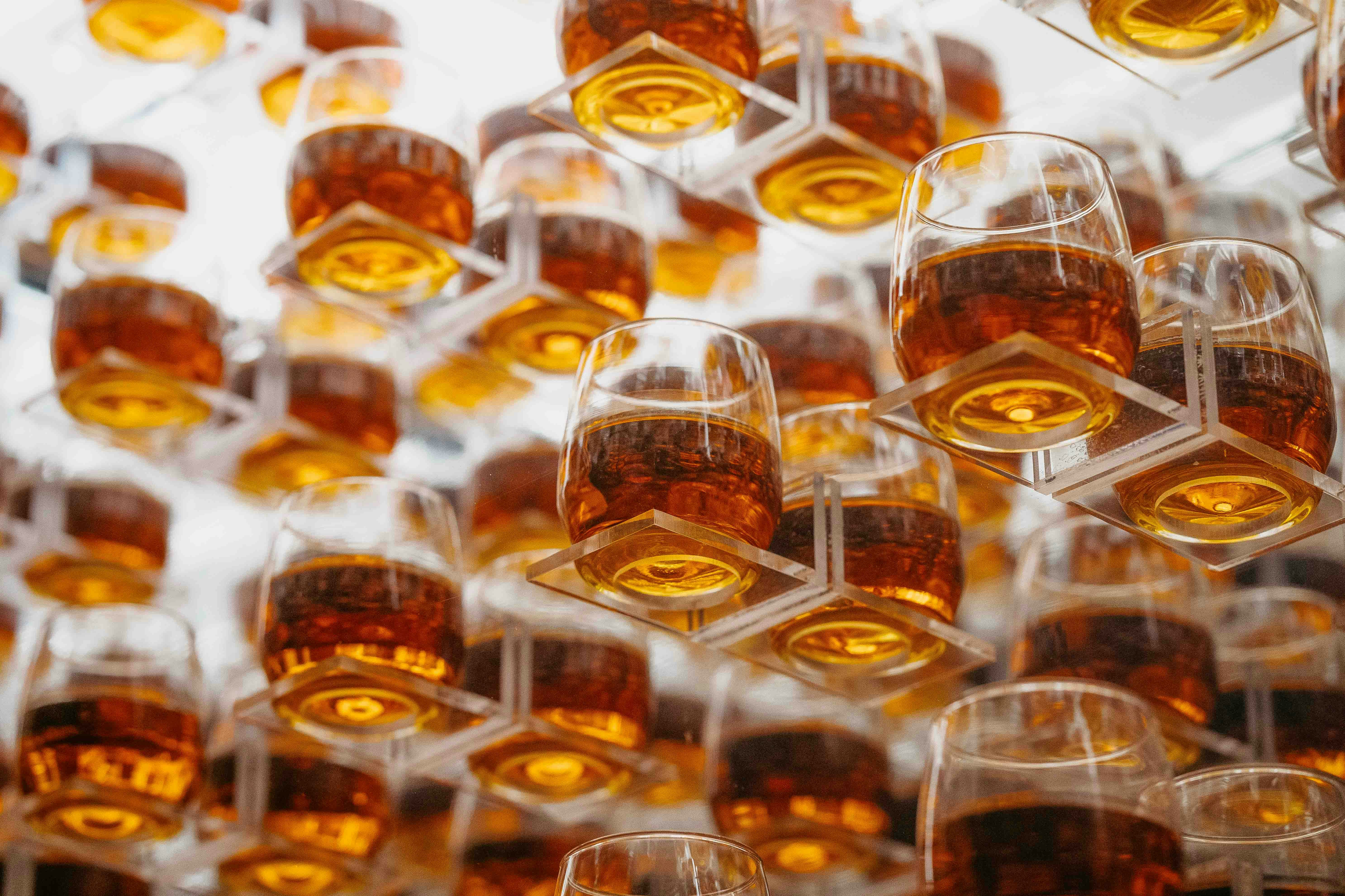 A collection of amber-colored whiskey glasses arranged in a geometric pattern, each glass resting on a clear, square platform, creating a visually striking display.