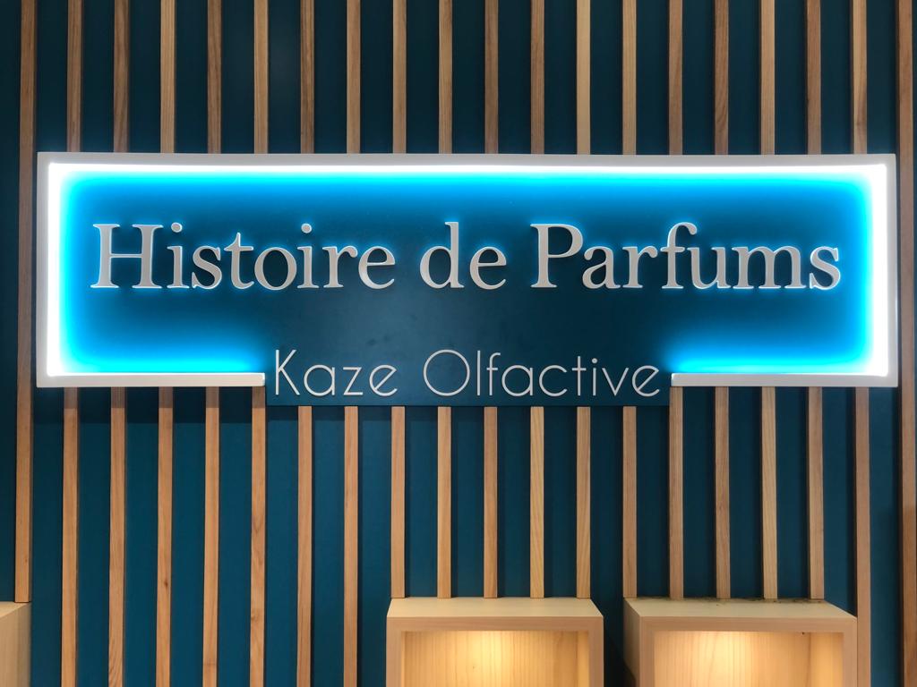 Une enseigne lumineuse affiche en lettres blanches « Histoire de Parfums, Kaze Olfactive ». Le fond est bleu, avec des lattes verticales en bois chaleureux, créant un look moderne et accueillant.
