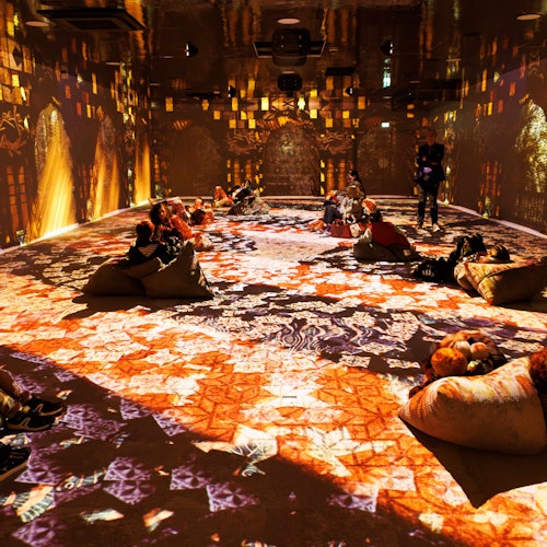 People sit on bean bags in a room illuminated by colorful, intricate light projections on the floors and walls.
