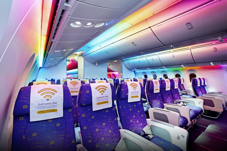 The interior of a modern airplane cabin featuring purple seats with white headrest covers displaying a Wi-Fi symbol. The cabin is illuminated with vibrant, multi-colored rainbow mood lighting along the ceiling and overhead bins.