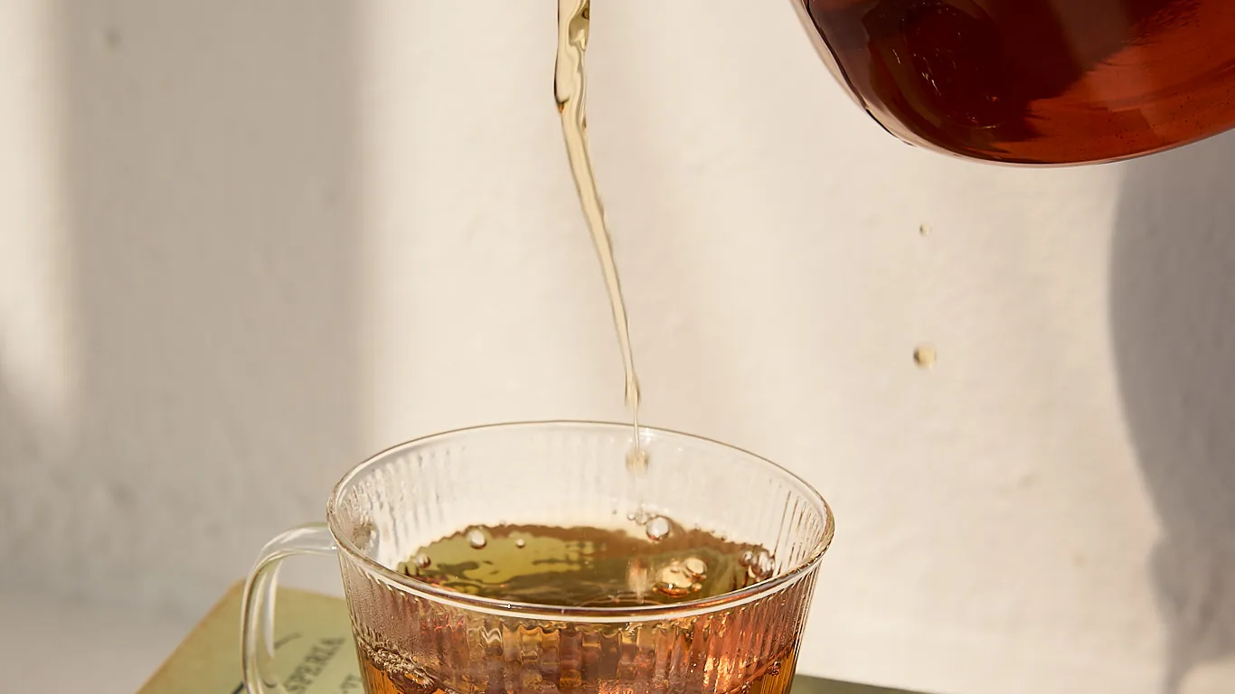 Sēkret tea poured from a glass teapot into a cup resting on a stack of books