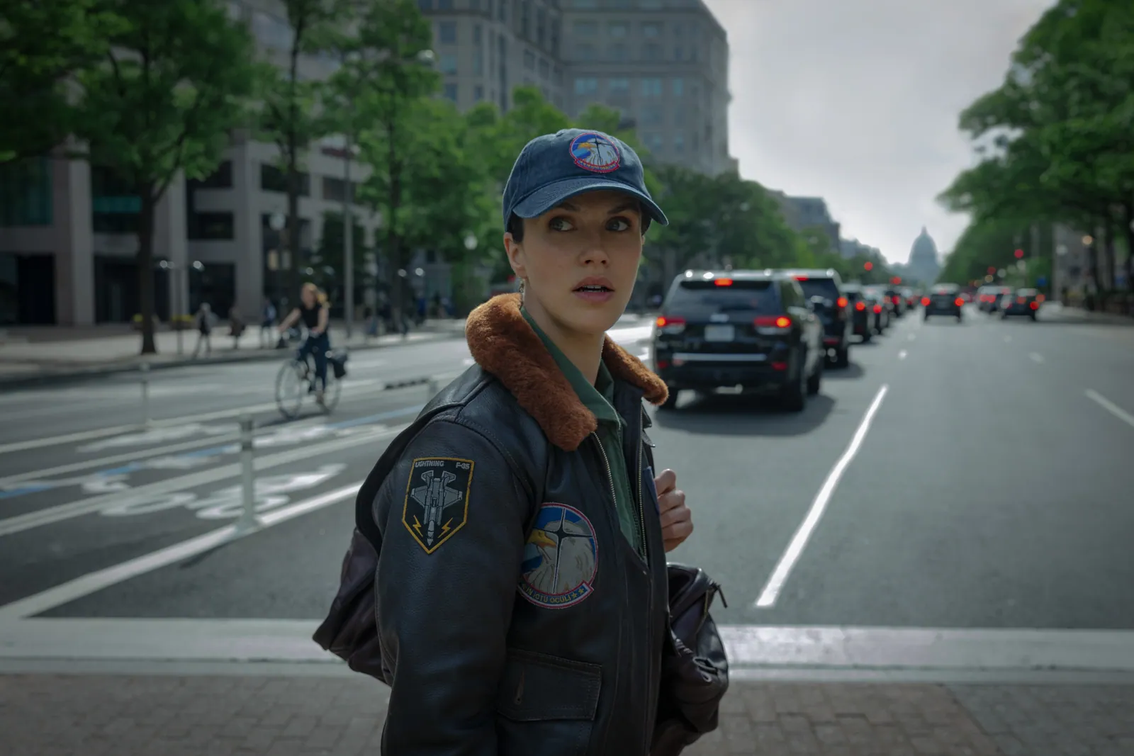 A person wearing a dark aviation jacket and cap stands on a city street with traffic and buildings in the background, creating an urban and dynamic atmosphere.