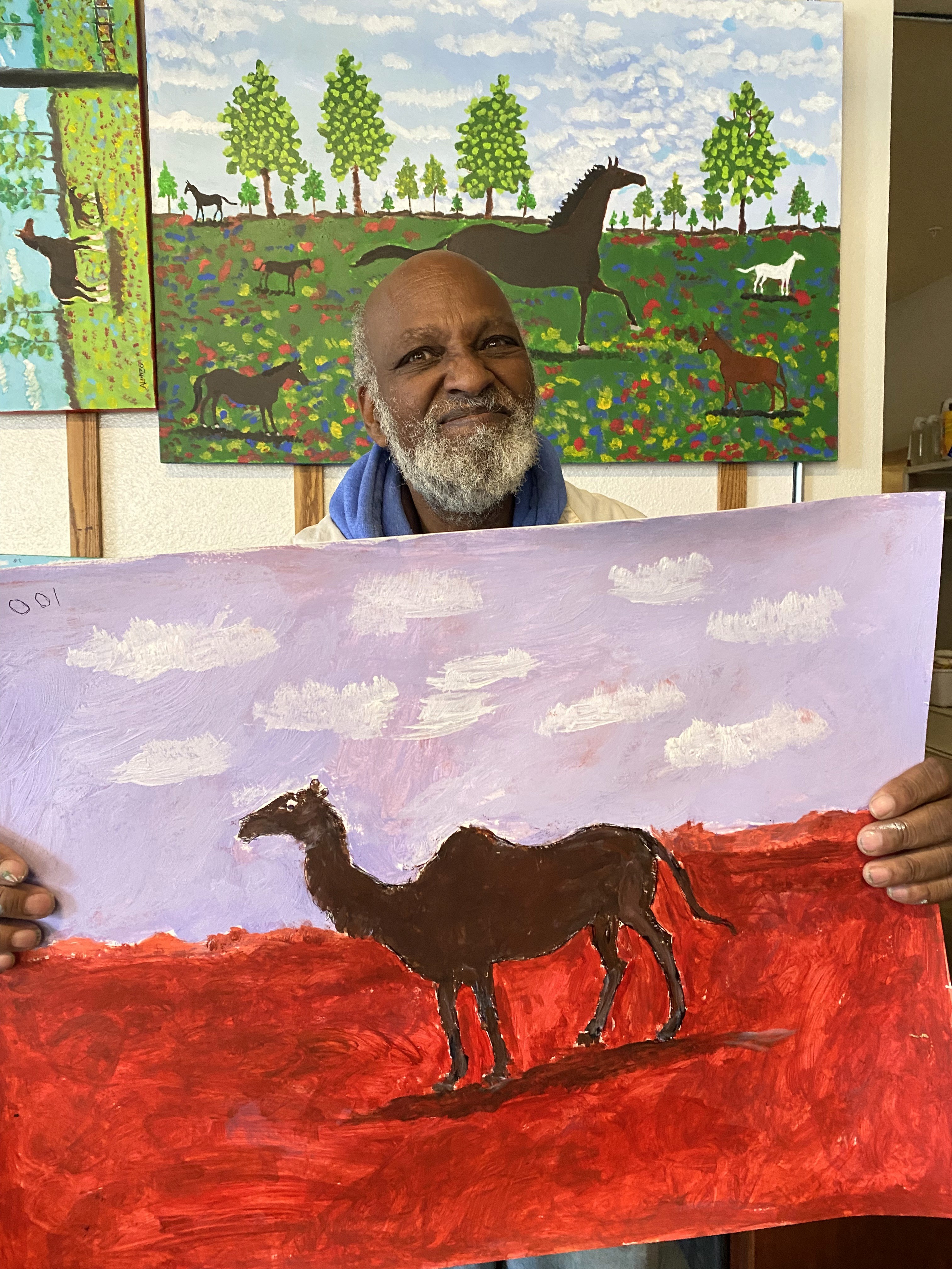 Portrait of Alonzo Clemons, Access Gallery artist, smiling in the studio while holding a brightly colored camel painting with horse painting displayed on wall behind him.