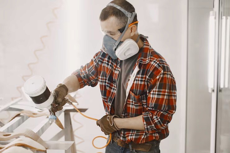 Man using paint sprayer, wearing a mask and goggles, staining wood. Wearing a plaid shirt and work gloves.