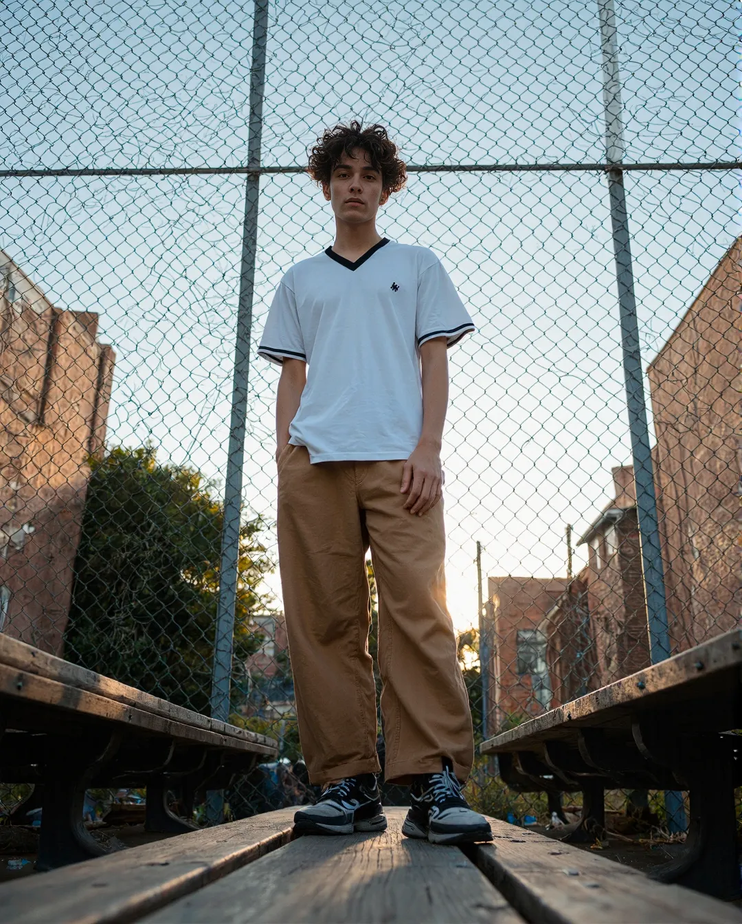 Fashion photograph on wooden walkway with chain-link fence and urban backdrop during golden hour, featuring casual white top and tan pants