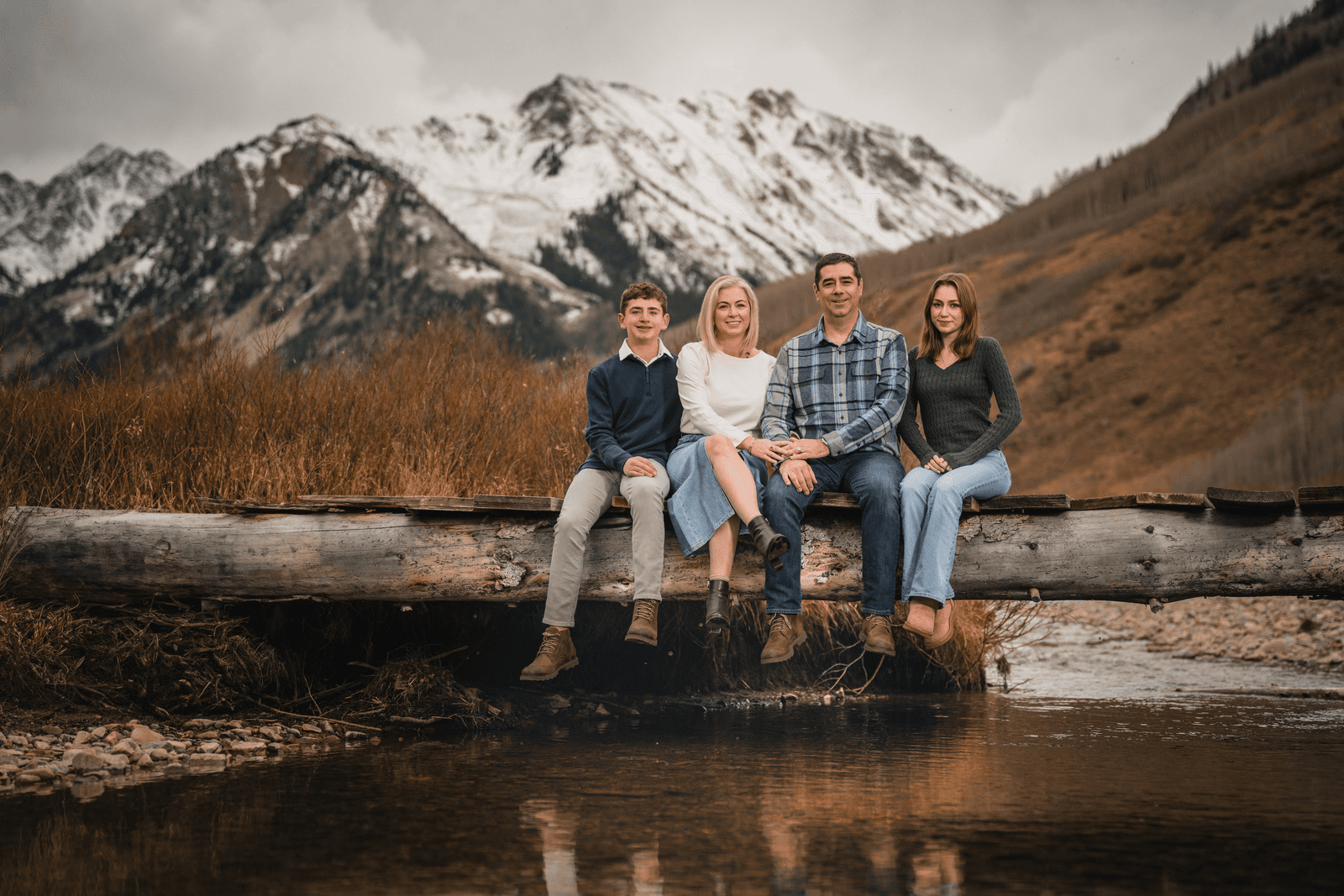 Four members of the Candes family sit on a fallen log over a creek, with brown autumn brush and snow-covered mountains behind them.