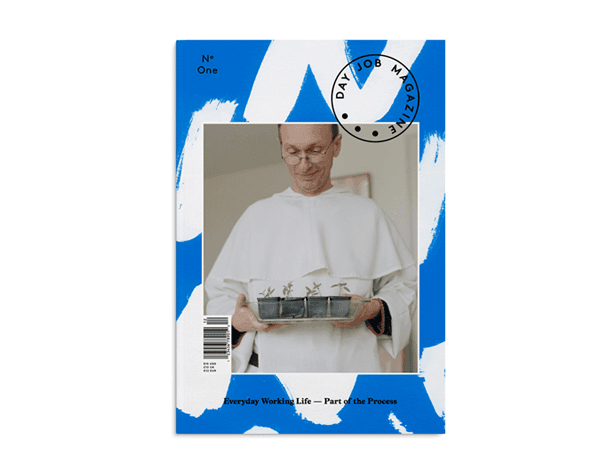 The image shows the cover of a magazine titled "Day Job Magazine". The cover is predominantly blue with white brushstroke-like patterns. A central photograph depicts a man wearing glasses and a white garment, holding a tray with four small containers. The magazine's issue number, "N° One", is visible in the top left corner. The cover text reads "Everyday Working Life — Part of the Process". A black circular stamp or seal appears in the top right corner. The magazine cover has a white border around the photograph.