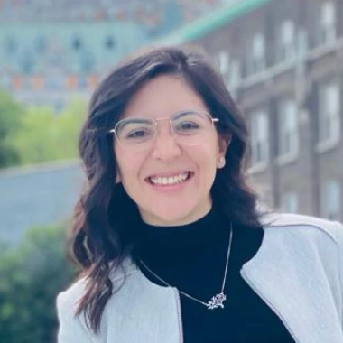 A smiling individual with glasses, wearing a light jacket over a black top, is standing outside with a blurred background of a building and some greenery.