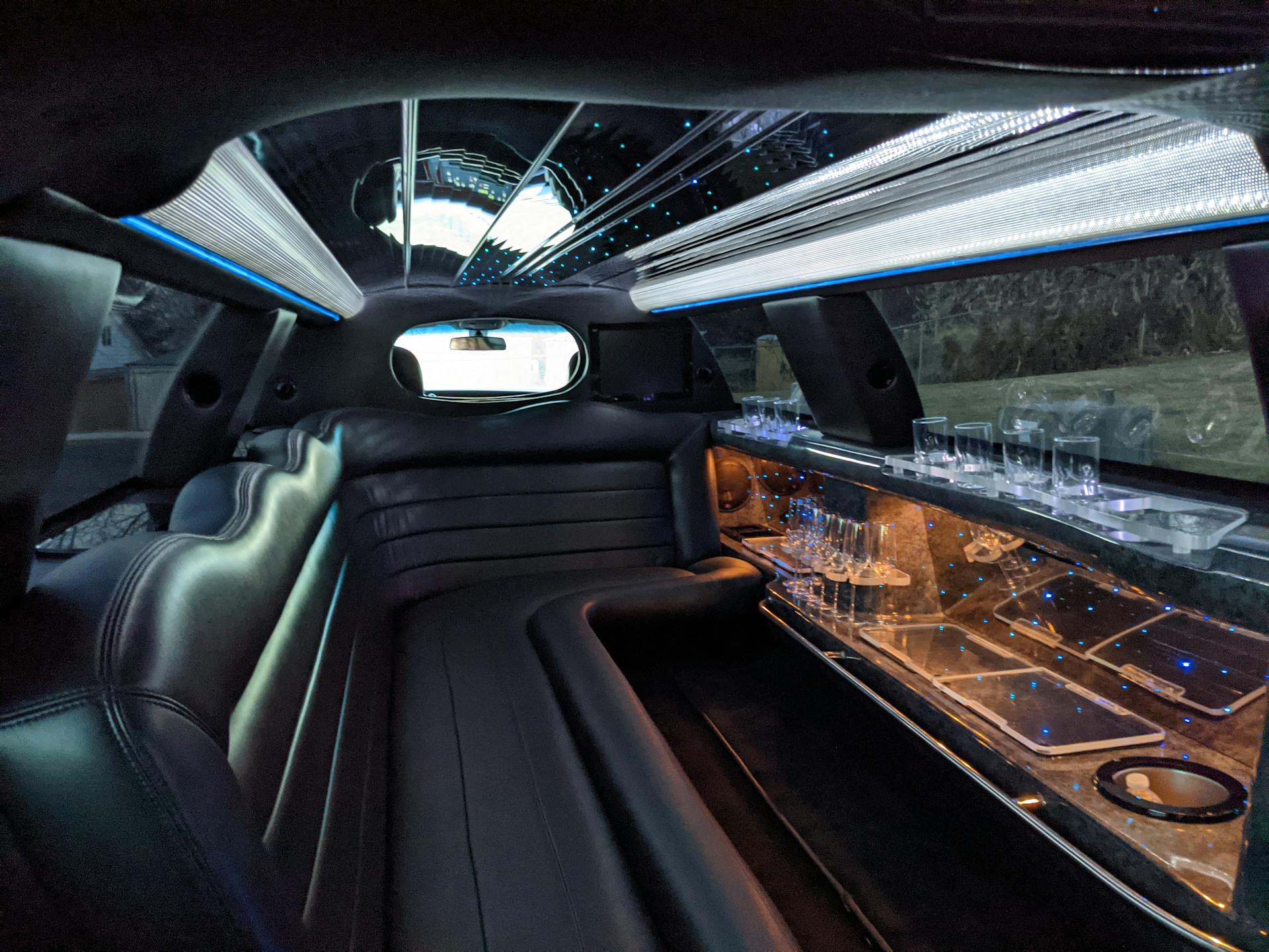 Interior of a luxury limousine featuring plush black leather seating, illuminated ceiling with star-like lights, and a bar area with glassware, emphasizing elegance for special occasion transportation services.