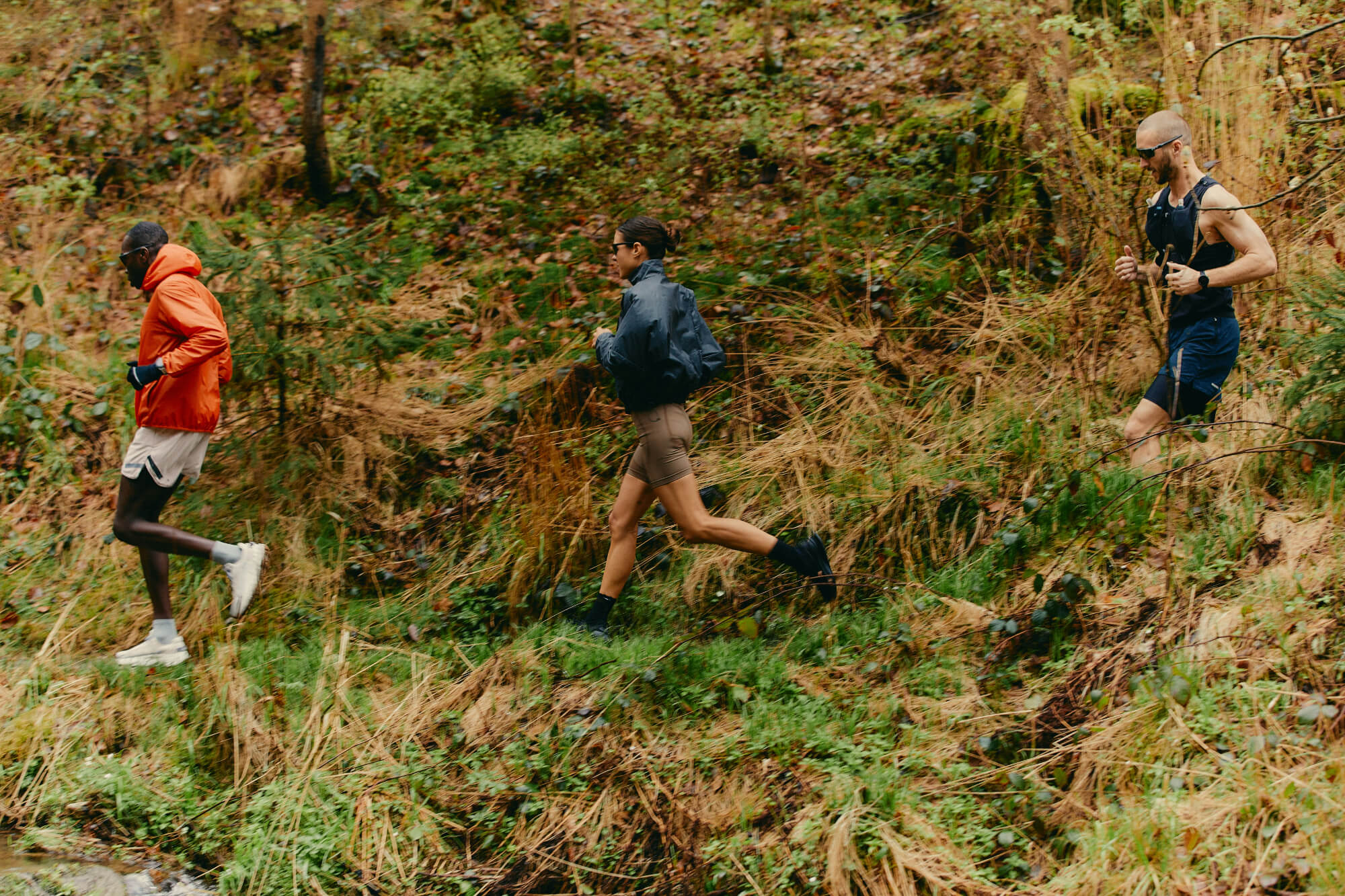 Three people jog energetically through a lush, green forest. One wears orange, another in a gray jacket, and the last in black, conveying determination and focus.