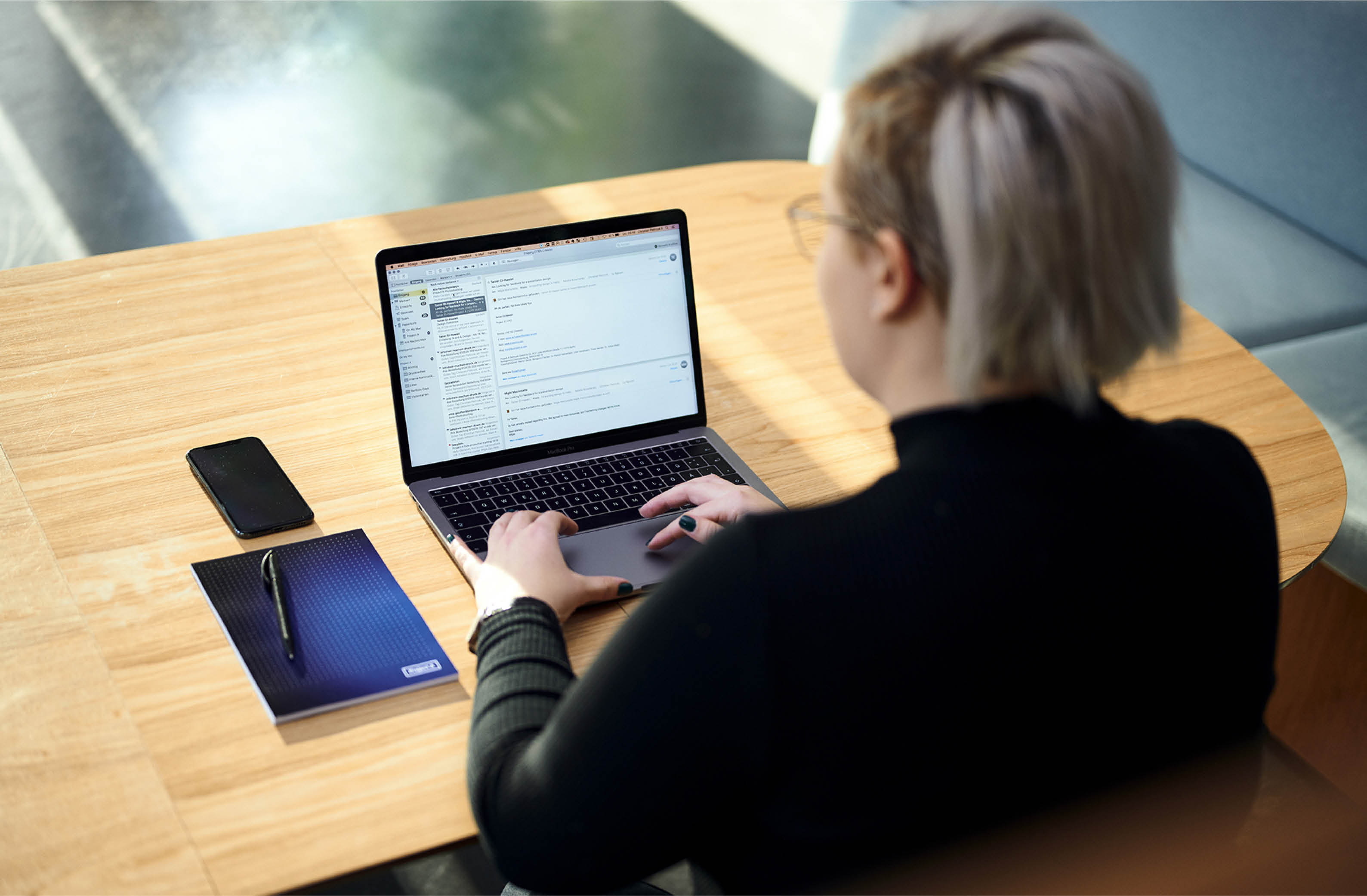 Project A employee working on a laptop at a wooden table with a branded notebook and phone, showcasing a productive workspace.