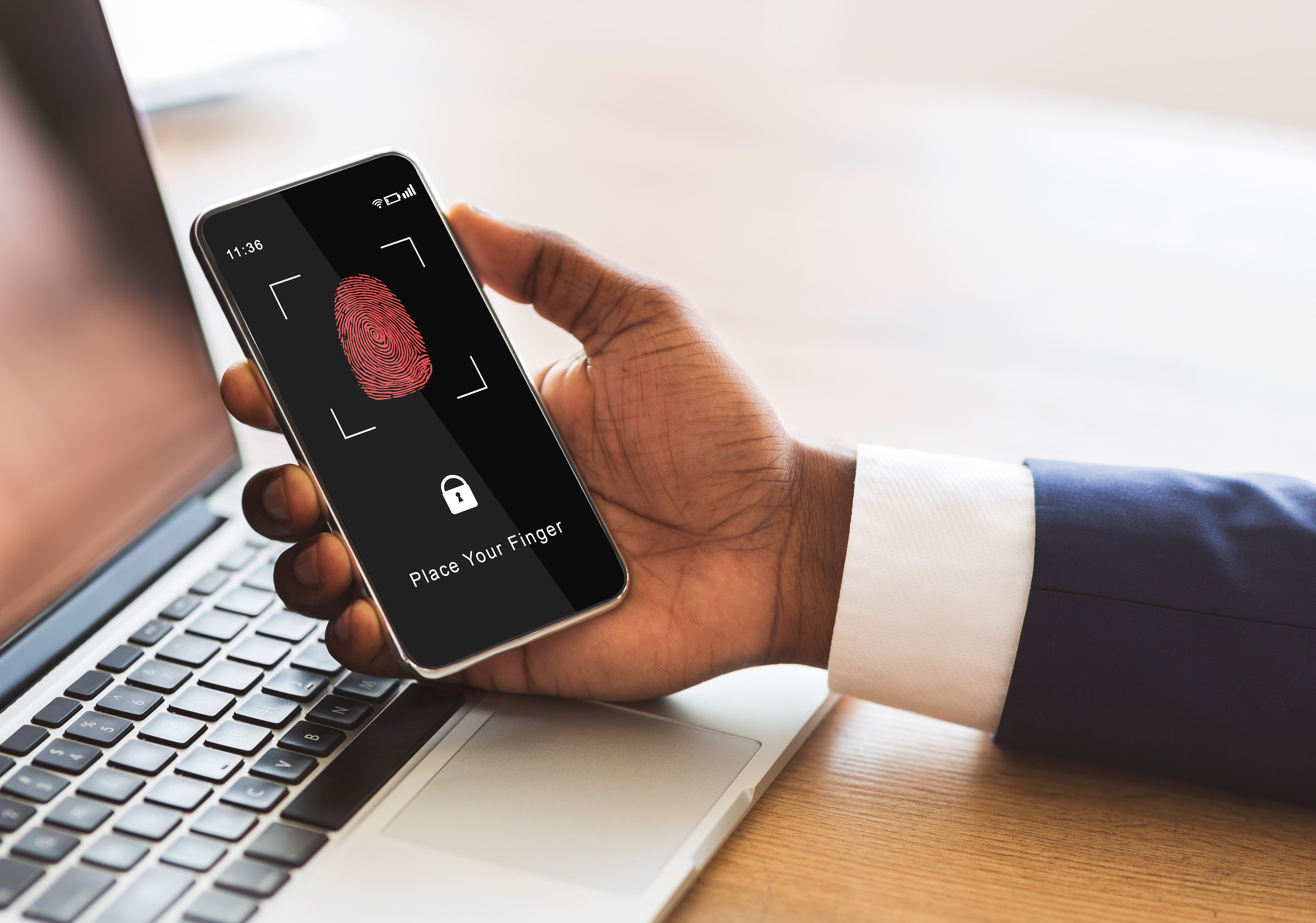 A close-up of an African American businessman holding a smartphone with a fingerprint authentication screen over a laptop, demonstrating cybersecurity and data protection.