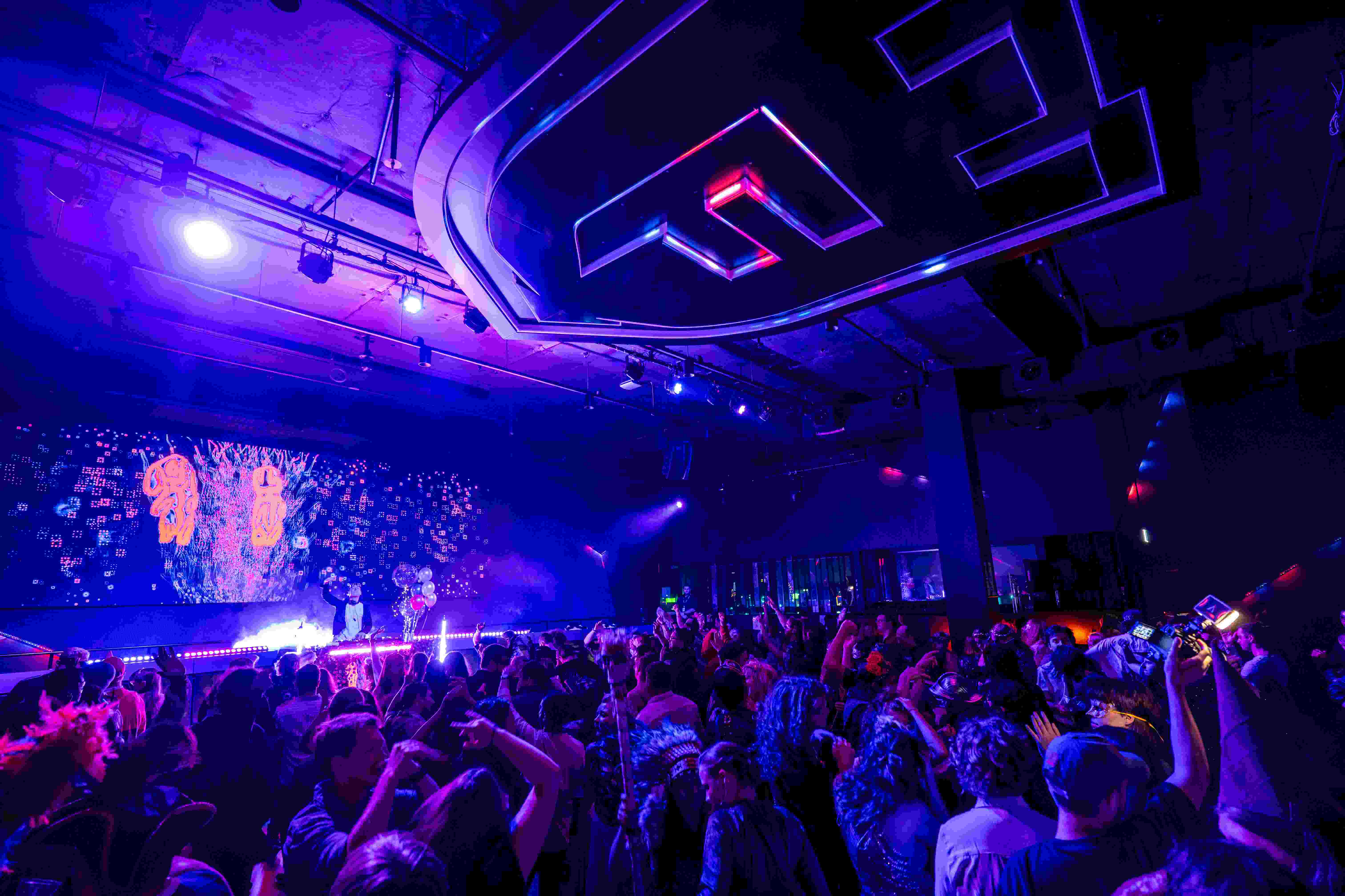 wide shot of nightclub under indigo lighting with throng of people dancing while DJ plays onstage