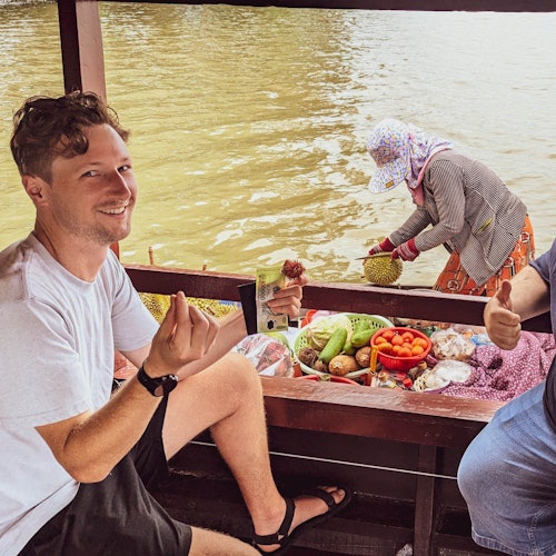 Dua pria tersenyum di atas perahu dengan berbagai buah dan sayuran; seseorang dengan topi dan masker sedang menangani hasil panen di perahu lain di dekatnya.