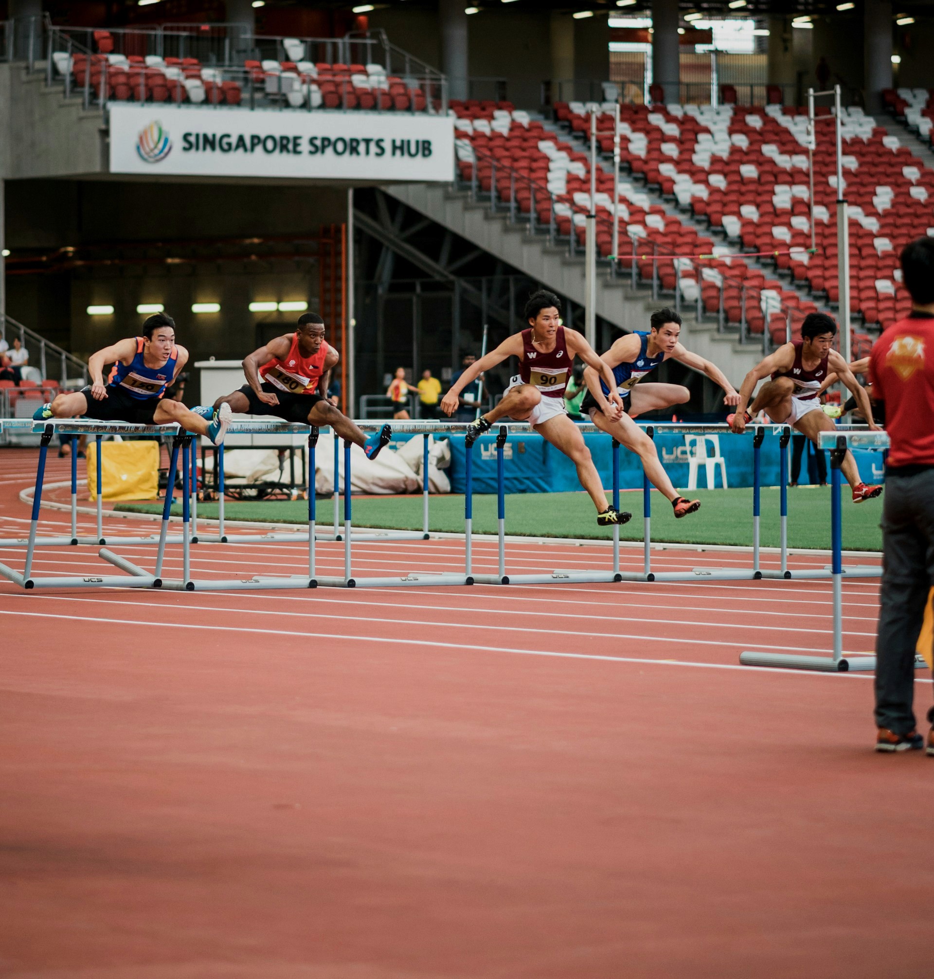 Four male athletes jump over hurdles during a track race at a stadium with red seats.