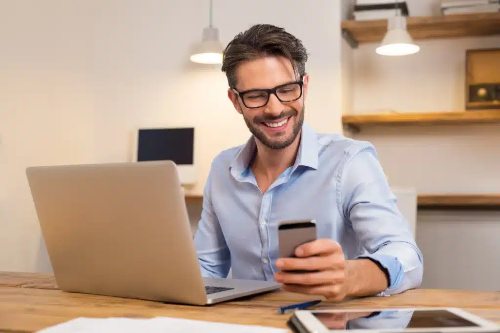 Top property management software professional smiling while checking property updates on smartphone beside laptop.