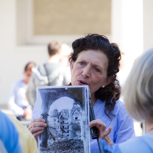 Nainen, joka pitelee mikrofonia ja esittelee mustavalkoista kuvaa vaurioituneesta rakennuksesta, taustalla epätarkkoja ihmisiä.