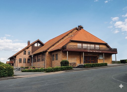 Außenansicht des Gemeindehauses der ECBG Detmold: Ein großes Backsteingebäude mit Satteldach und Holzbalkon unter blauem Himmel.