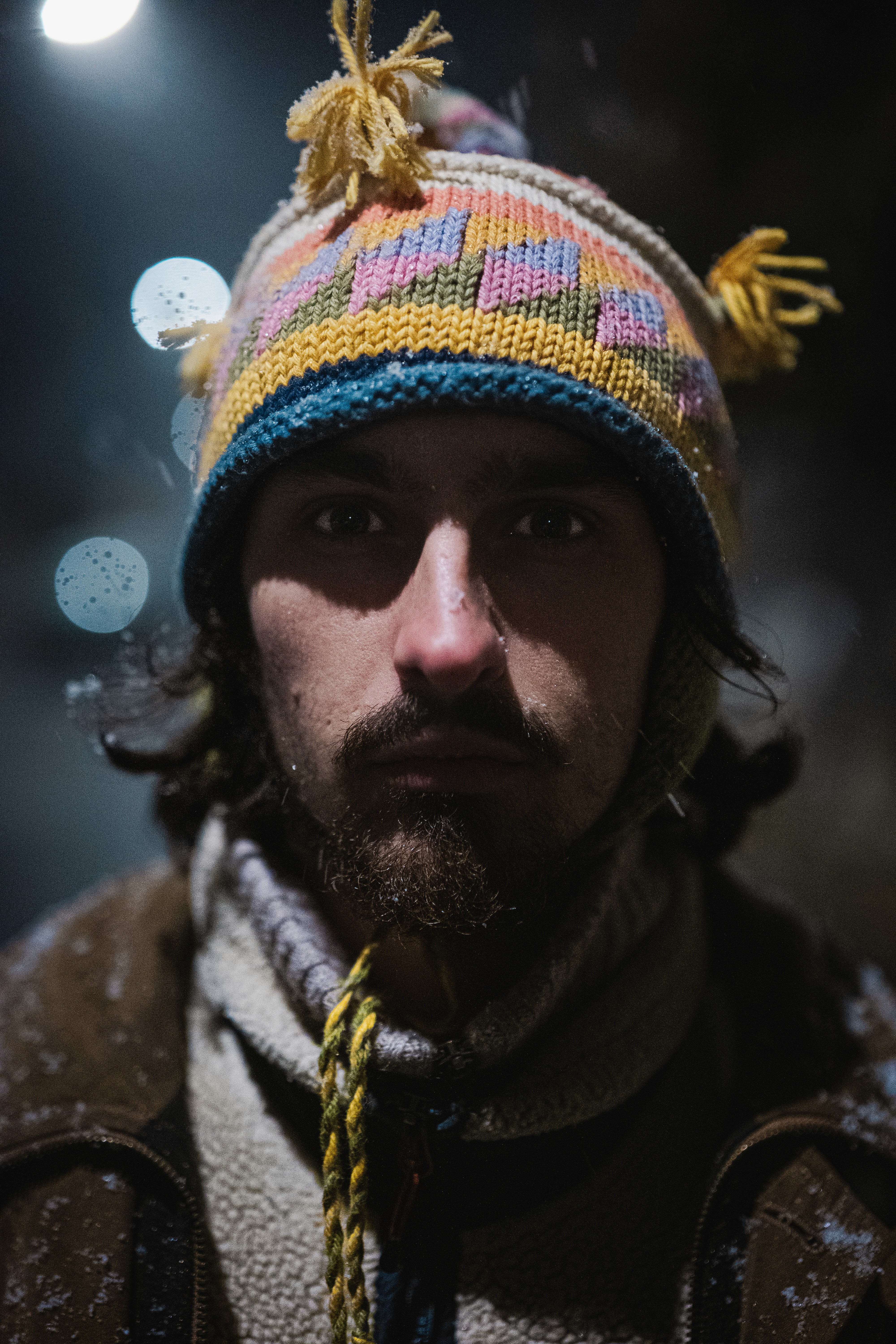A young person with brown hair, wearing reflective silver sports sunglasses and a black sweater, looks at the camera while resting their chin on their hand.