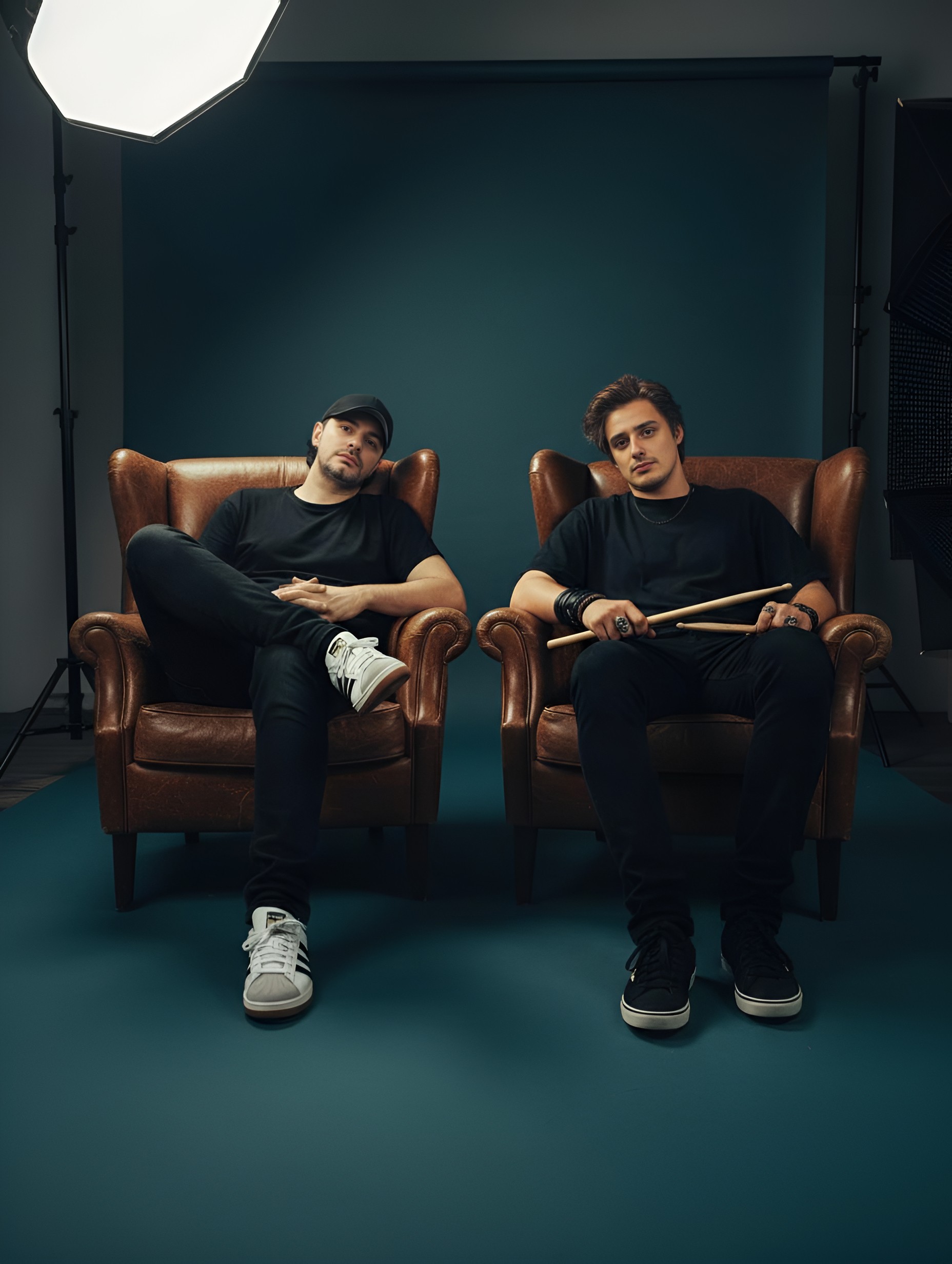 Two men in black shirts sitting on brown leather chairs in a studio, one holding drumsticks.