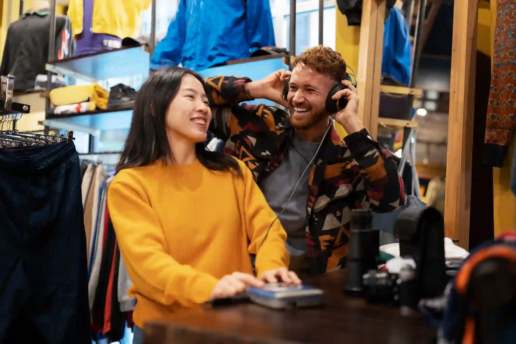 Two people in a store, smiling while one listens to music on headphones, suggesting a fun shopping experience.
