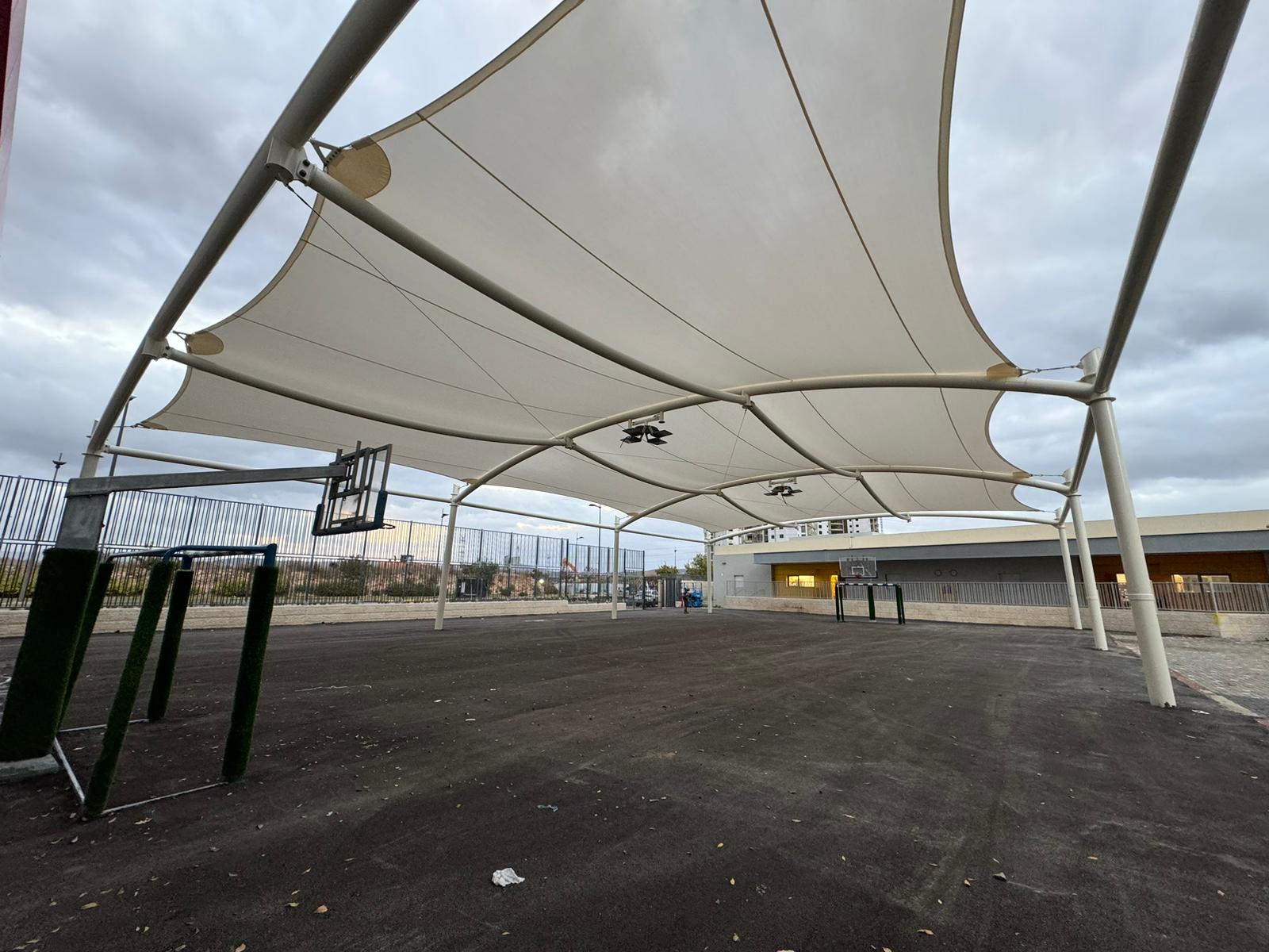 Covered outdoor area with basketball hoop and paved ground under a large beige shade sail supported by a steel frame.