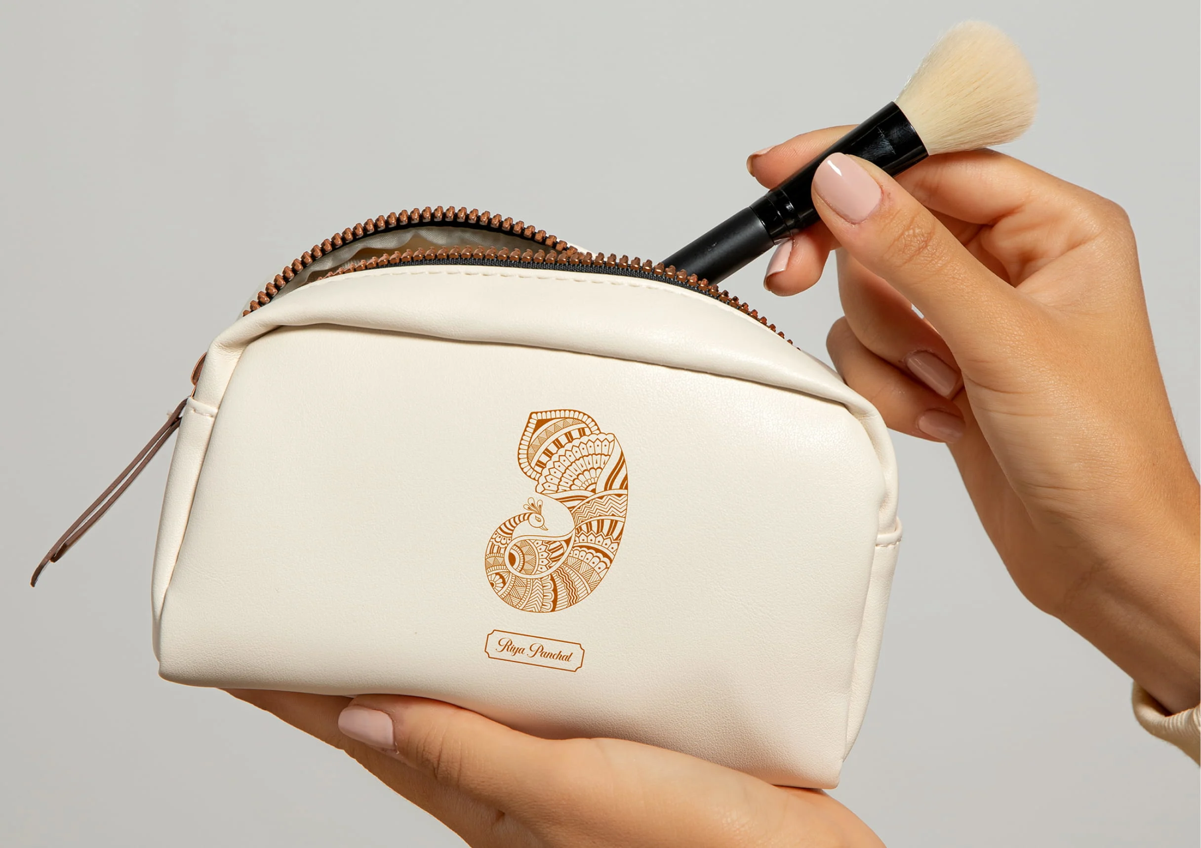 Hands hold a cream cosmetic pouch with a decorative logo while a makeup brush rests above it, highlighting beauty and skincare essentials.