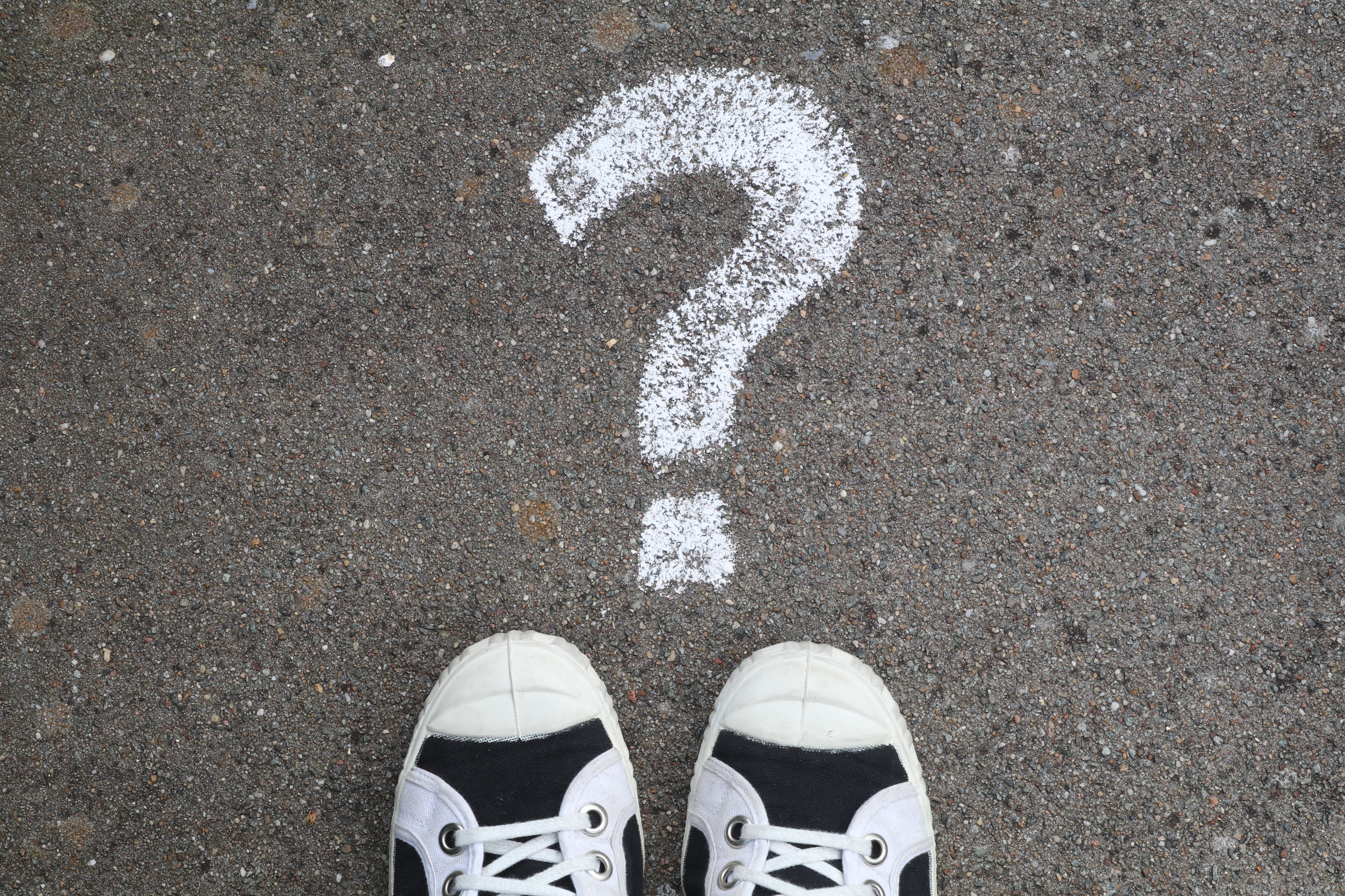 A pair of black and white sneakers is positioned on a gray asphalt pavement facing a large white chalk question mark drawn on the ground, symbolizing uncertainty or inquiry.