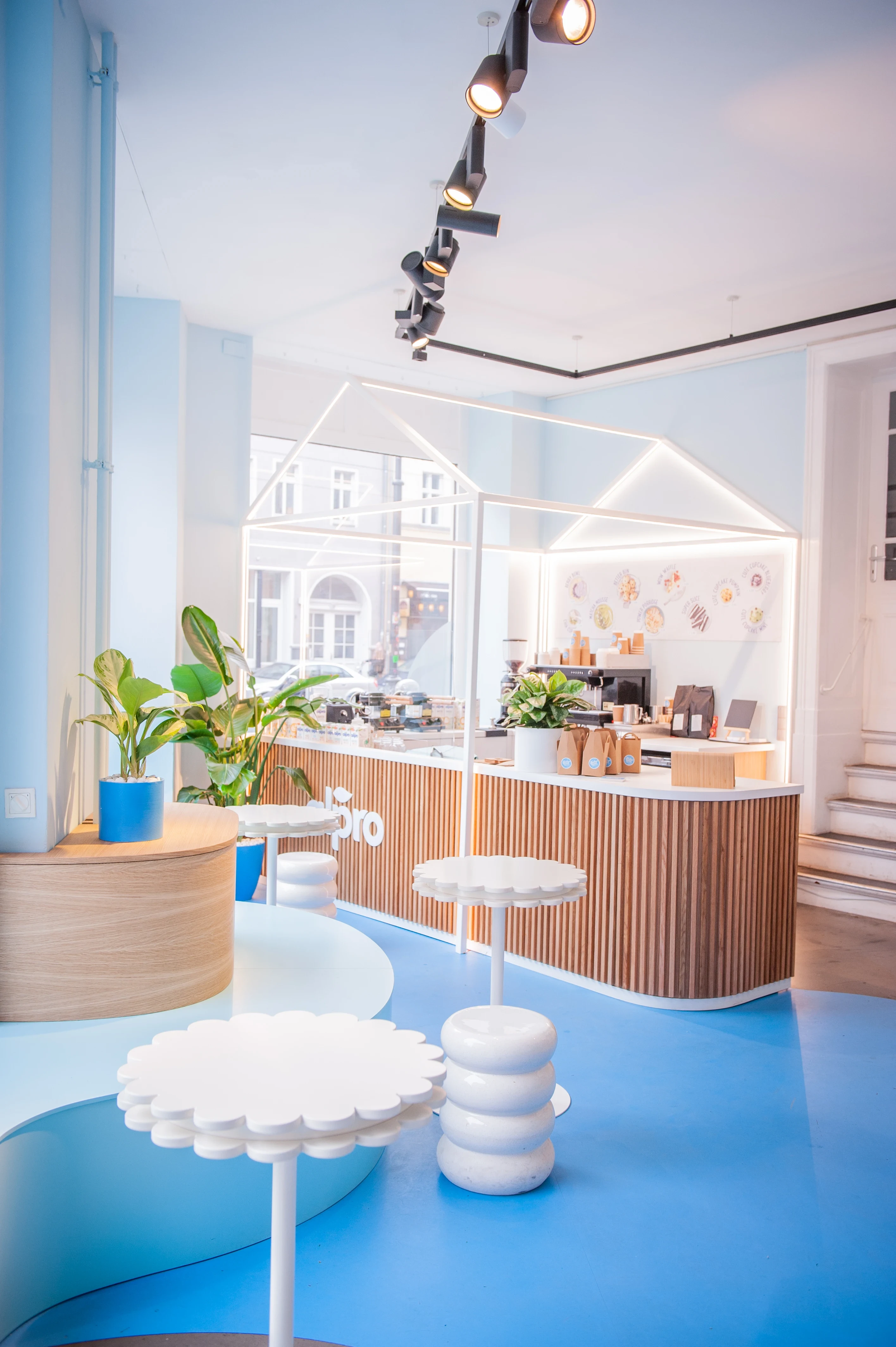 Cafe counter framed by a glowing house outline, with flower tables and ribbed stools on the blue floor of the Alpro pop-up.