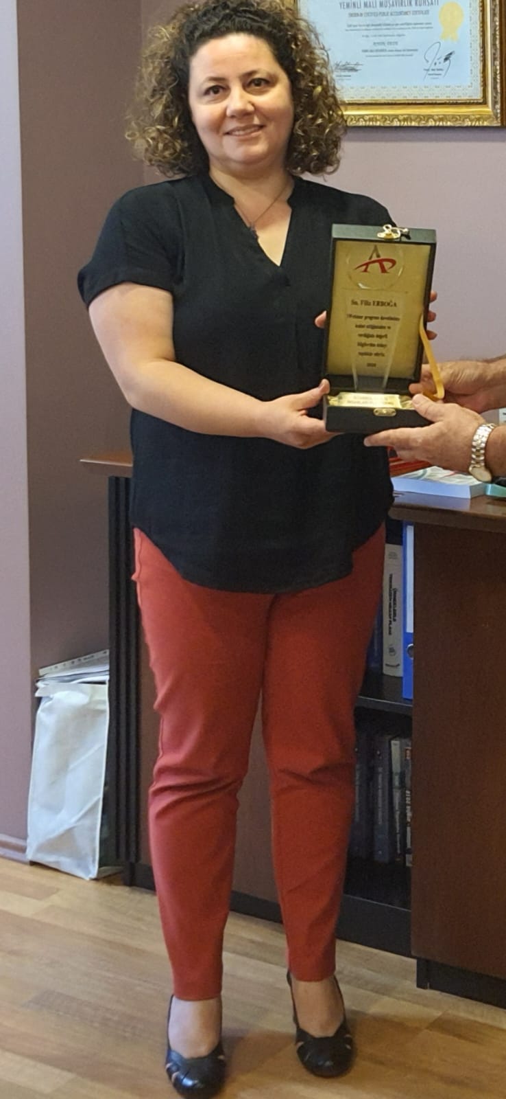 A smiling woman with her arms crossed, standing against a dark green background. She has long, dark hair.