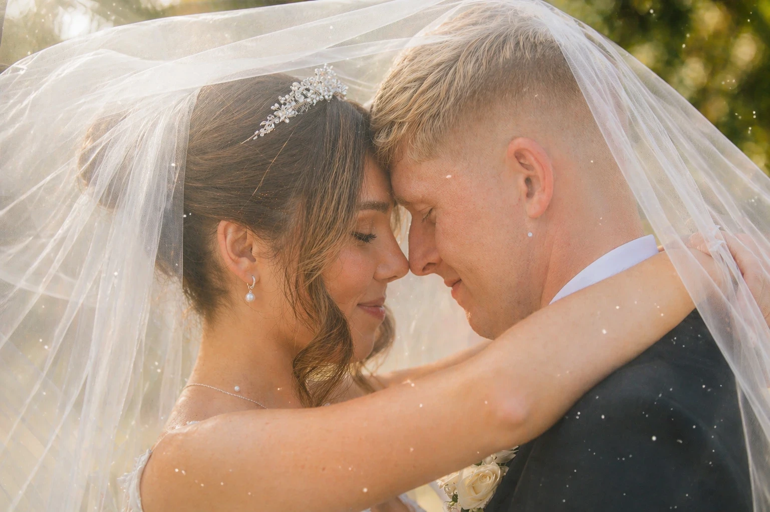 Bride & groom embrace under her veil. Eyes closed, smiling gently. Tiara, pearl earrings & necklace.