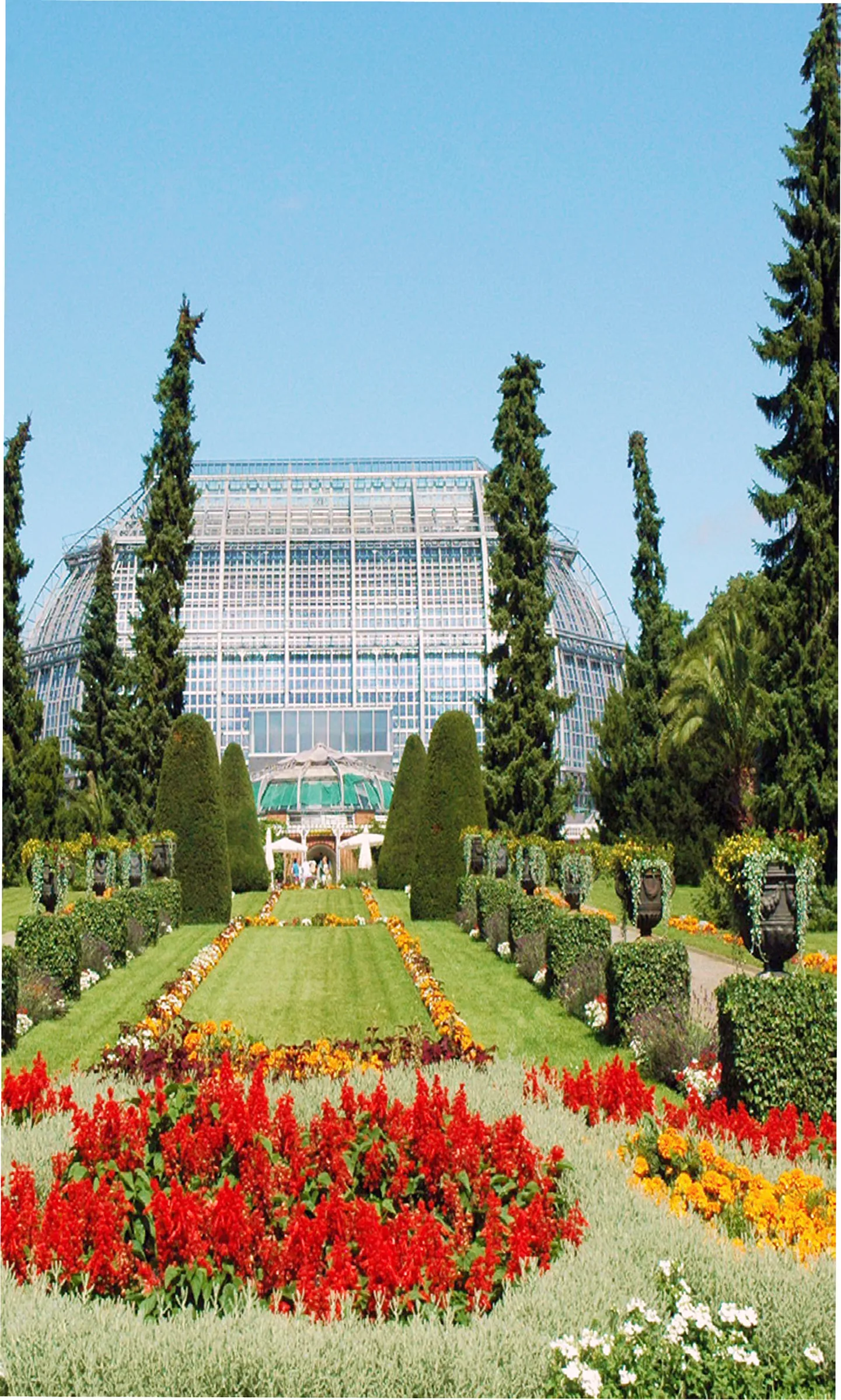 Das Große Tropenhaus im Botanischen Garten Berlin an einem sonnigen Tag, im Vordergrund bunte Blumenbeete und Rasenflächen.