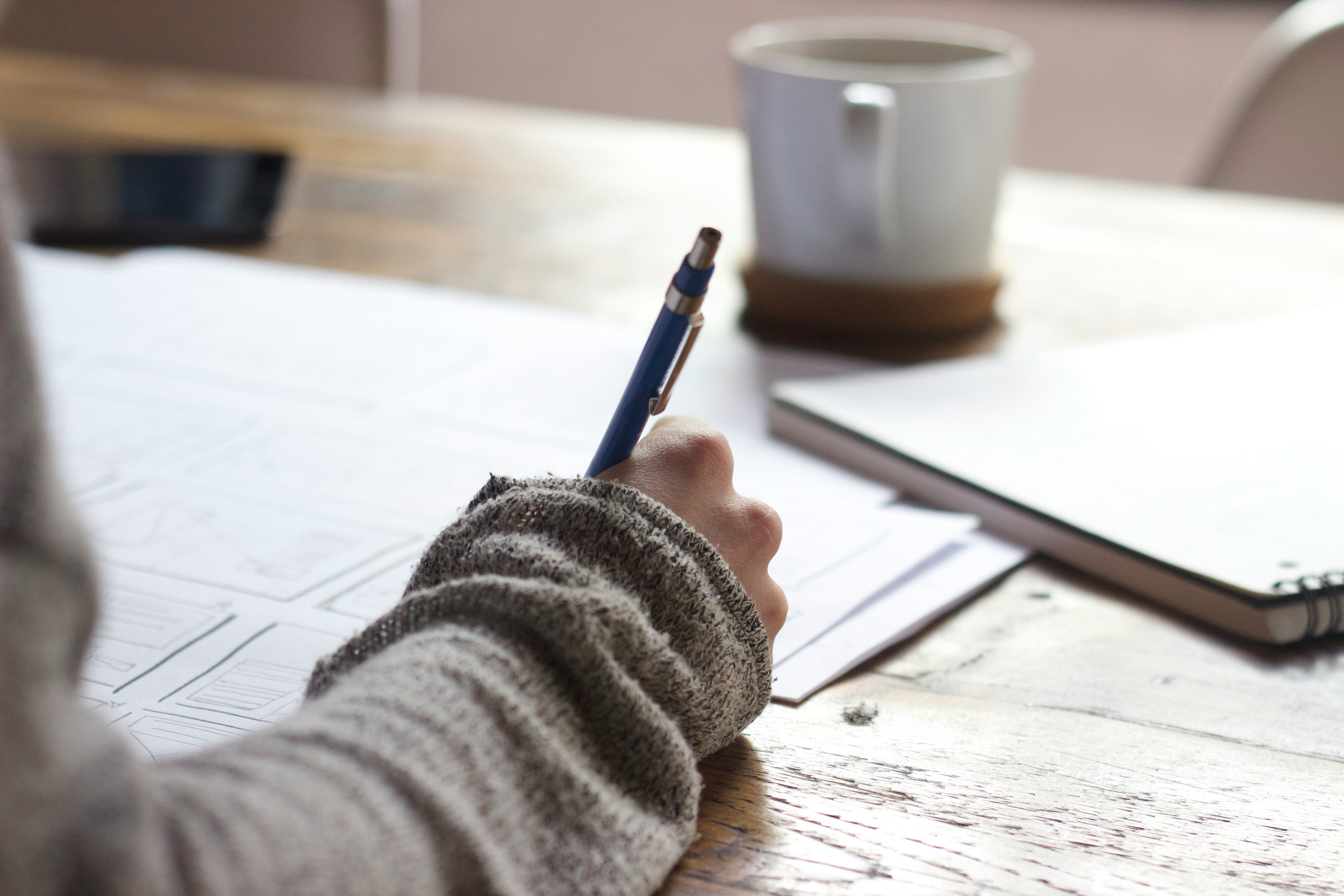 An arm lies atop several pieces of paper on a desk. The person holds a pen and is writing. In the background white mug sits on the desk.