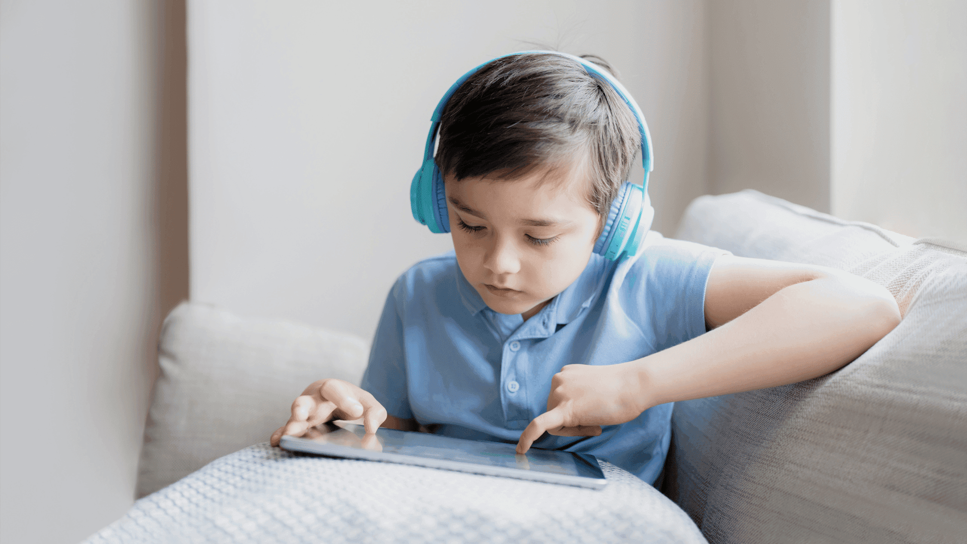 A young boy sitting on a sofa, wearing a blue polo shirt and blue headphones, holding a tablet while listening to HushAway®’s Sound Sanctuary.