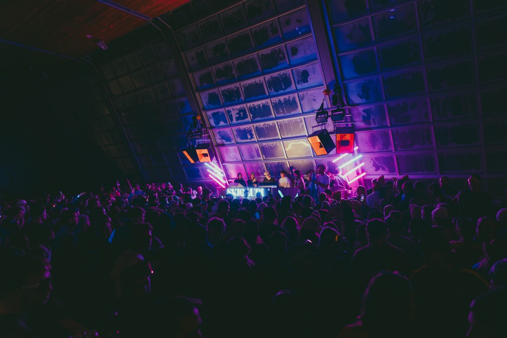 Crowded music event with DJ and vibrant lights.
