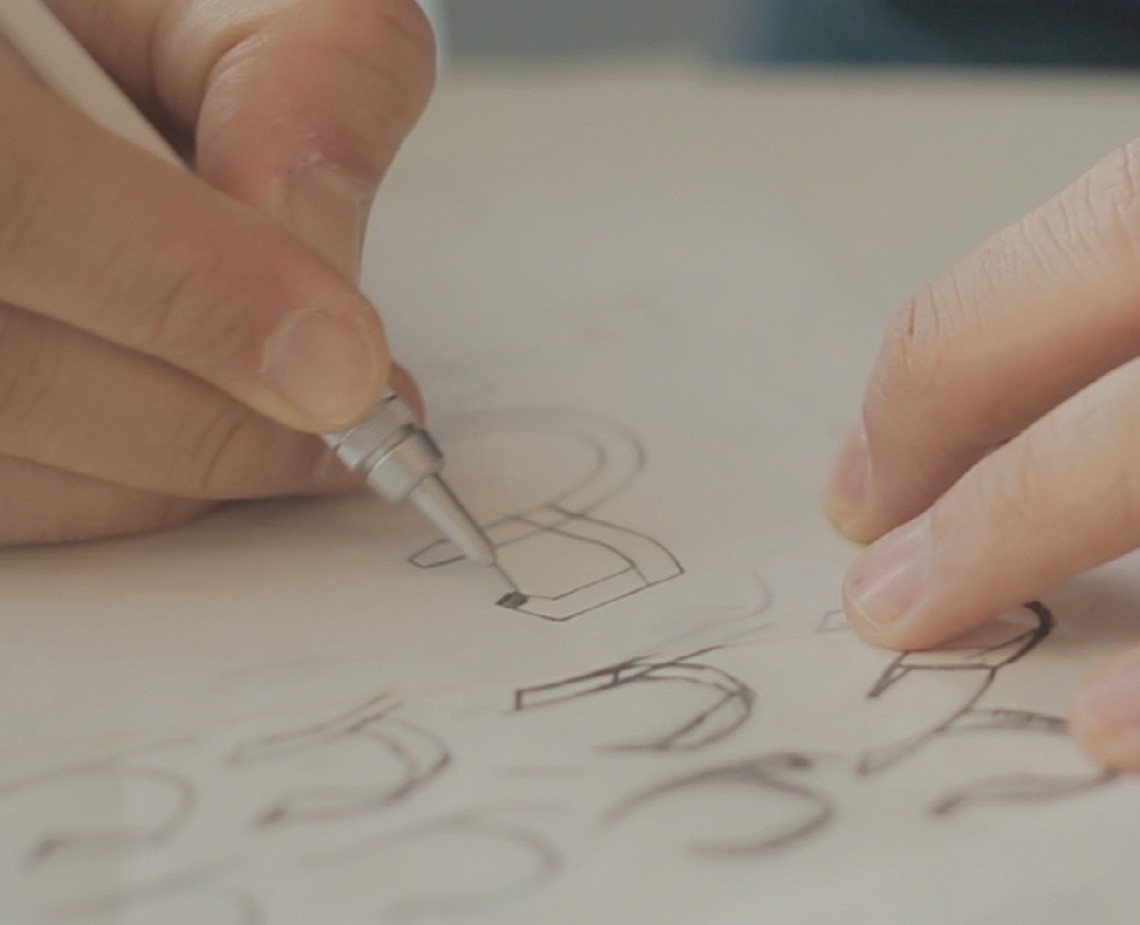 Two hands are shown drawing on a white sheet of paper. The left hand holds a silver pen and is drawing a curved line. The right hand rests on the paper near the drawing. The drawing consists of curved lines and shapes resembling a design or pattern.