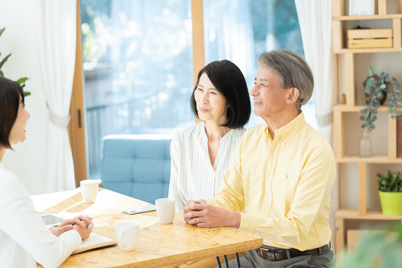 明るいリビングのテーブルで、シニア世代の夫婦が向かい側に座るスタッフと笑顔で話し合っている様子。